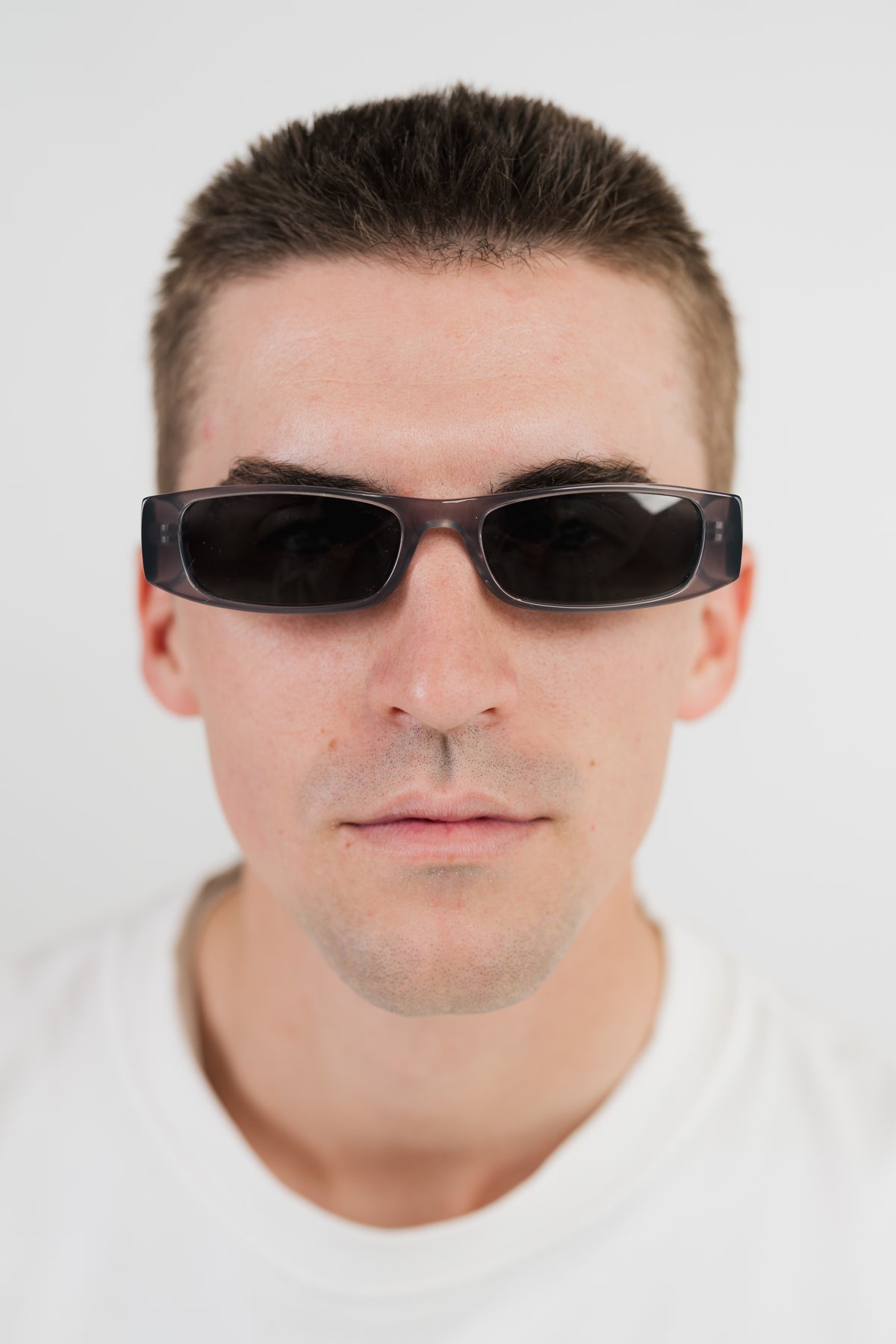 A close-up portrait of a man wearing dark rectangular sunglasses with a glossy black frame and dark lenses, set against a plain light gray background.
