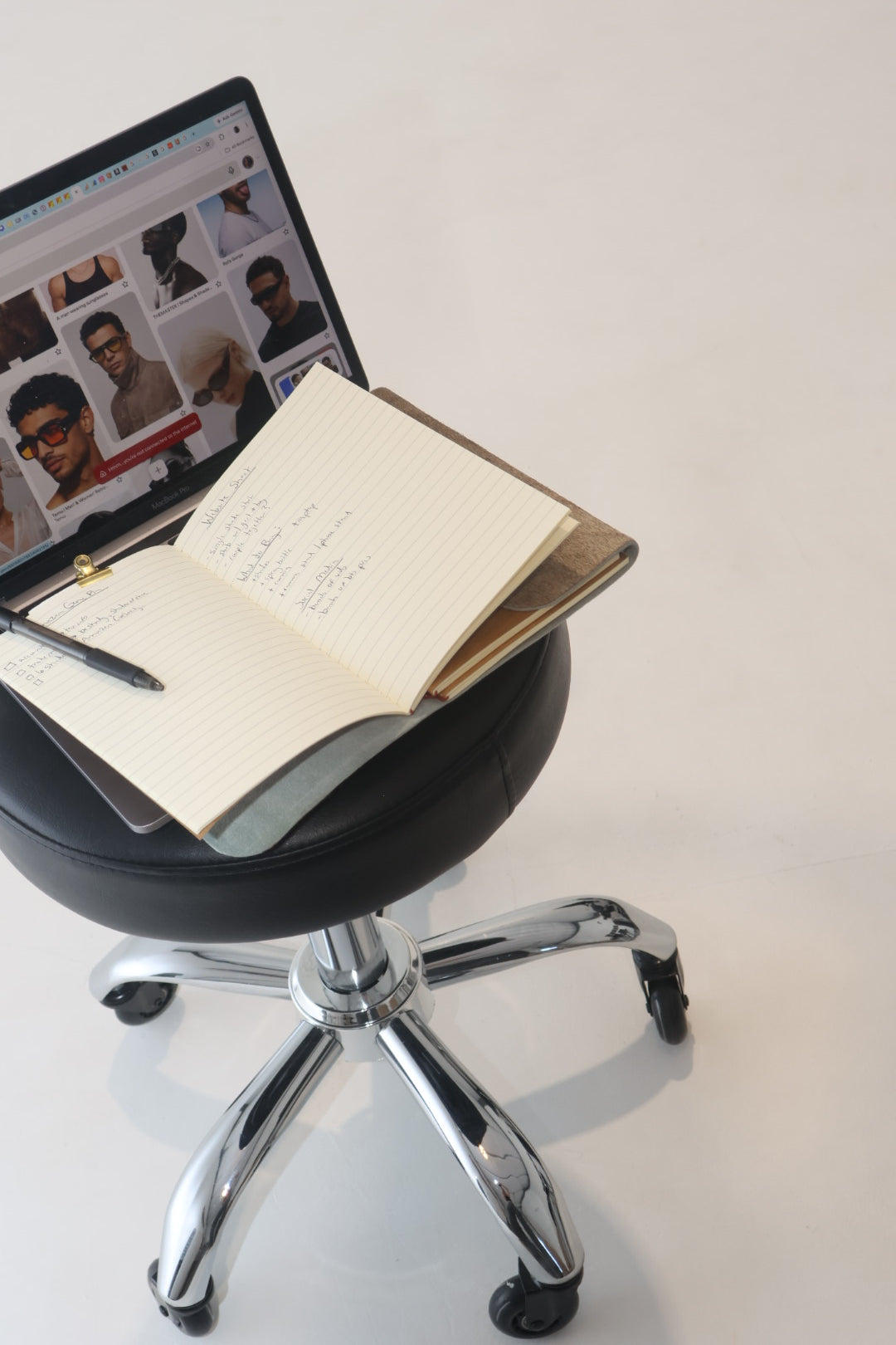 An open notebook with handwritten notes and a pen rests on a black swivel stool, with a laptop showing a website in the background against a plain light wall.