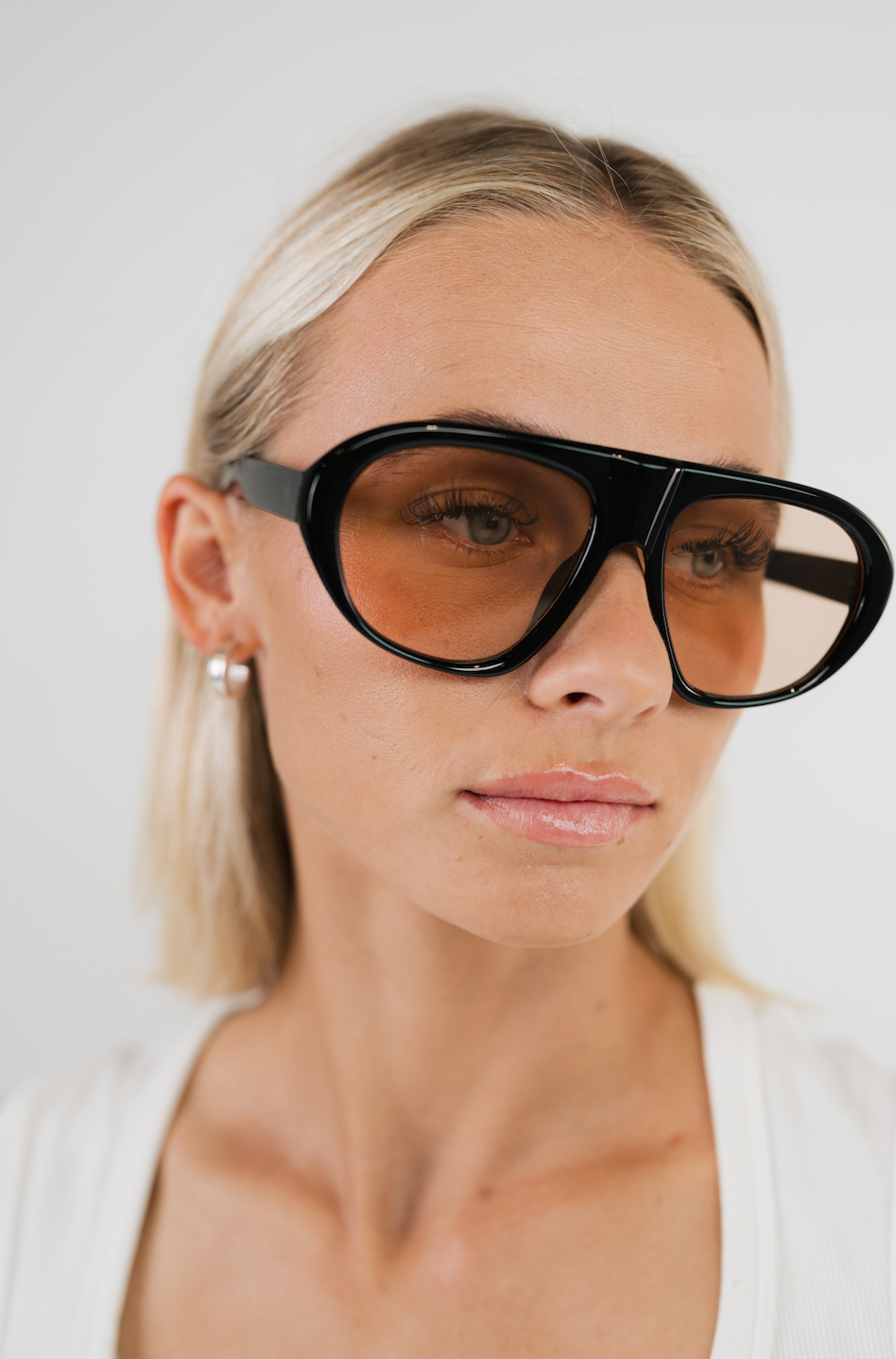 A close-up of a model wearing oversized square sunglasses with a thick black frame and amber-tinted lenses, paired with a white top against a light gray background.