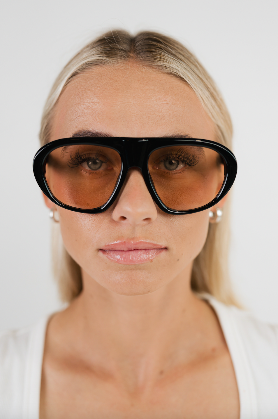 A close-up of a woman with blonde hair wearing oversized black sunglasses with tinted amber lenses and small pearl earrings, against a plain light background.