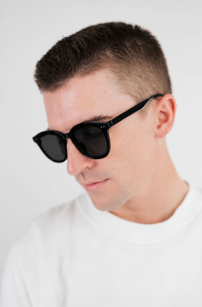 A close-up shot of a man wearing black sunglasses with a white crewneck shirt, photographed against a clean white background.