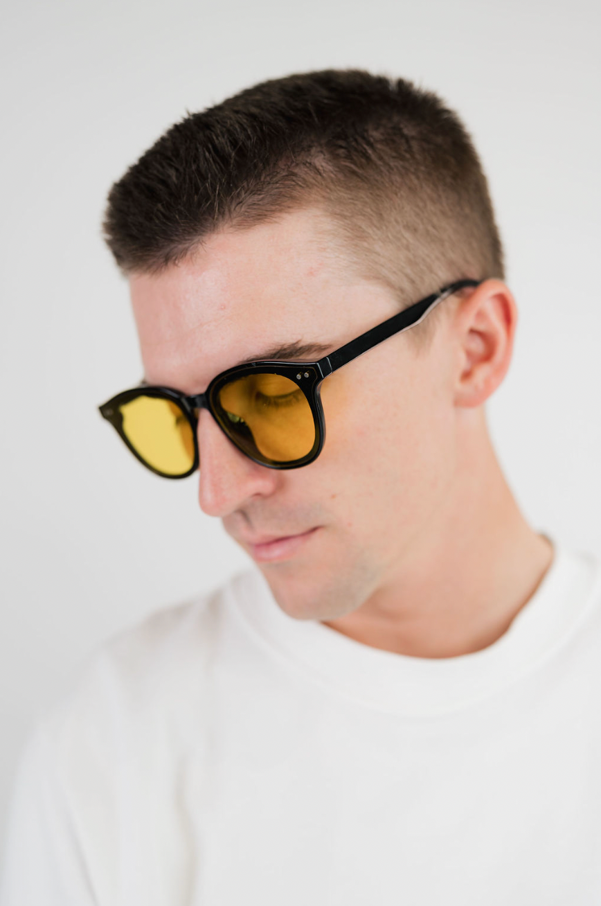 A close-up of a man wearing black-framed sunglasses with yellow tinted lenses, looking down and to the side against a plain white background.