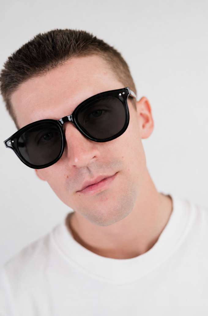 A close-up shot of a man wearing black sunglasses with a thick frame and dark lenses, dressed in a white crewneck t-shirt against a plain light gray background.