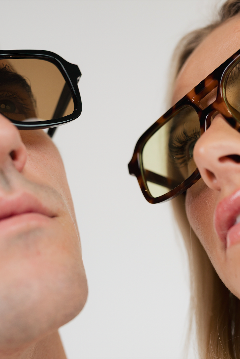 Close-up of two people wearing oversized square sunglasses, one with a black frame and brown tinted lenses and the other with a tortoiseshell frame and yellow-tinted lenses, against a plain light background.