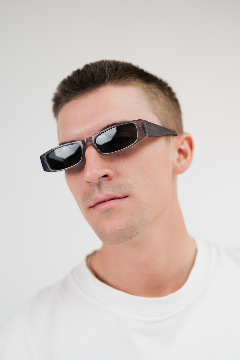 A man wearing futuristic angular black sunglasses with a small rectangular device on the temple, photographed against a clean light gray background.