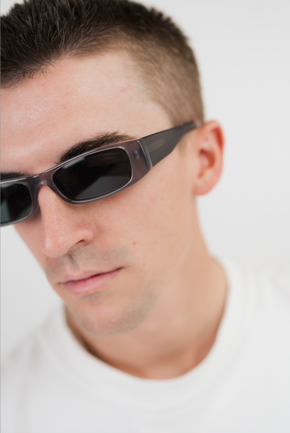 A close-up shot of a man wearing dark gray rectangular sunglasses with black lenses and a white t-shirt against a plain white background.