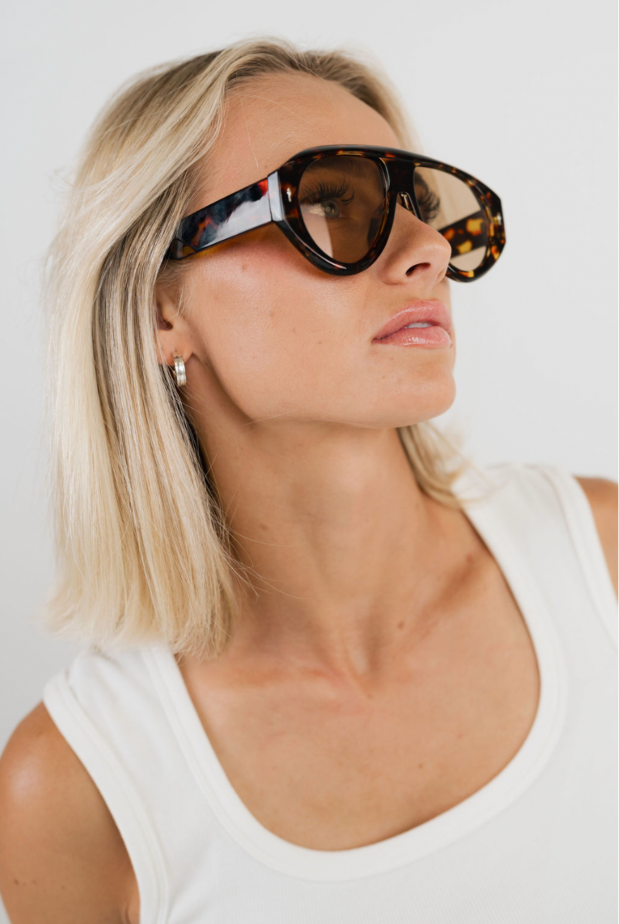 A close-up of a blonde woman wearing oversized tortoiseshell sunglasses with dark brown lenses and a white ribbed tank top, photographed against a clean white background.