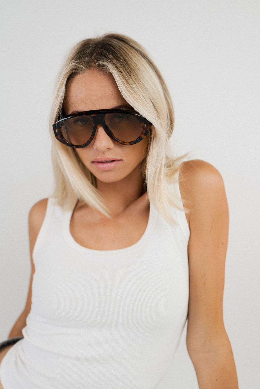 A blonde woman wearing oversized dark sunglasses and a white tank top, photographed against a plain light background.