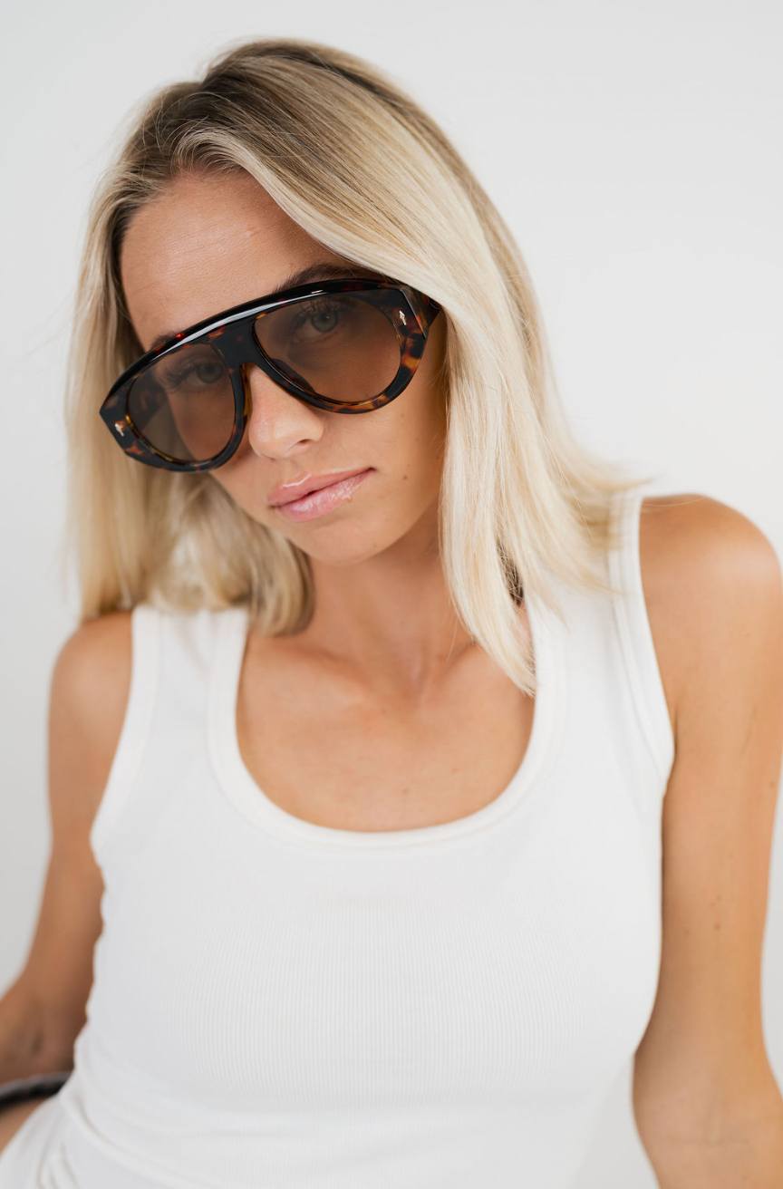 A blonde woman wearing oversized tortoiseshell sunglasses and a white ribbed tank top, photographed against a clean white background.