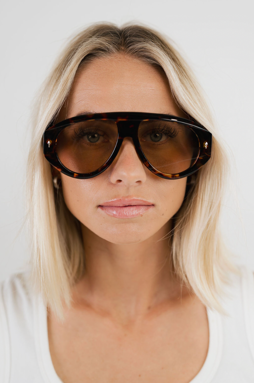 A close-up of a blonde woman wearing oversized tortoiseshell aviator-style sunglasses with dark brown tinted lenses and a white top against a plain light background.