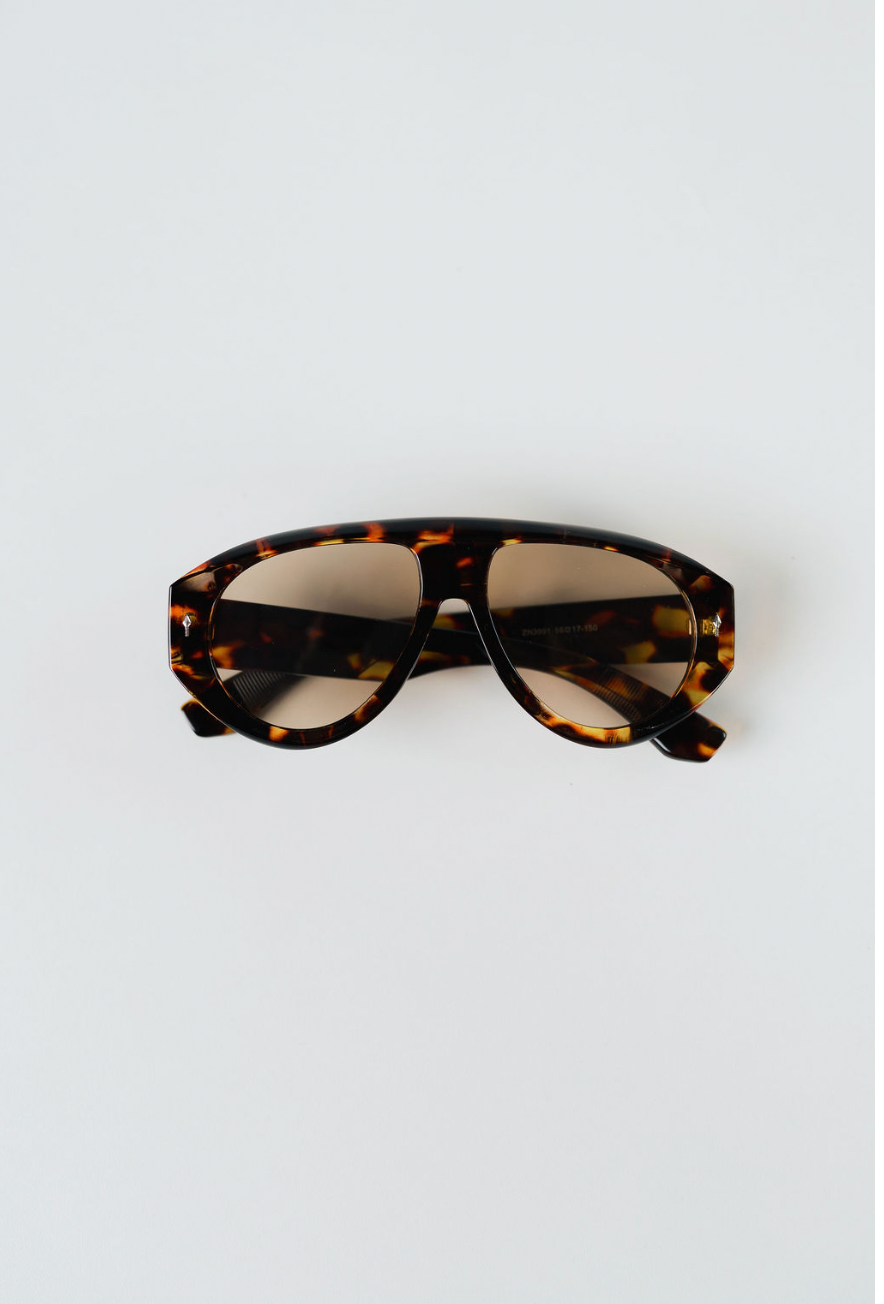 Tortoiseshell aviator-style sunglasses with a thick brown and black patterned frame and tinted lenses, photographed from above on a light gray background.