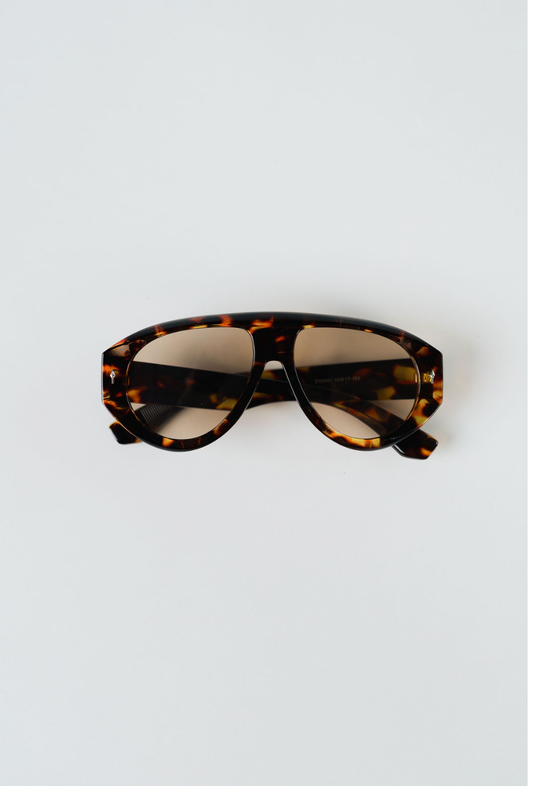 Tortoiseshell aviator-style sunglasses with a thick brown and black patterned frame and tinted lenses, photographed from above on a light gray background.