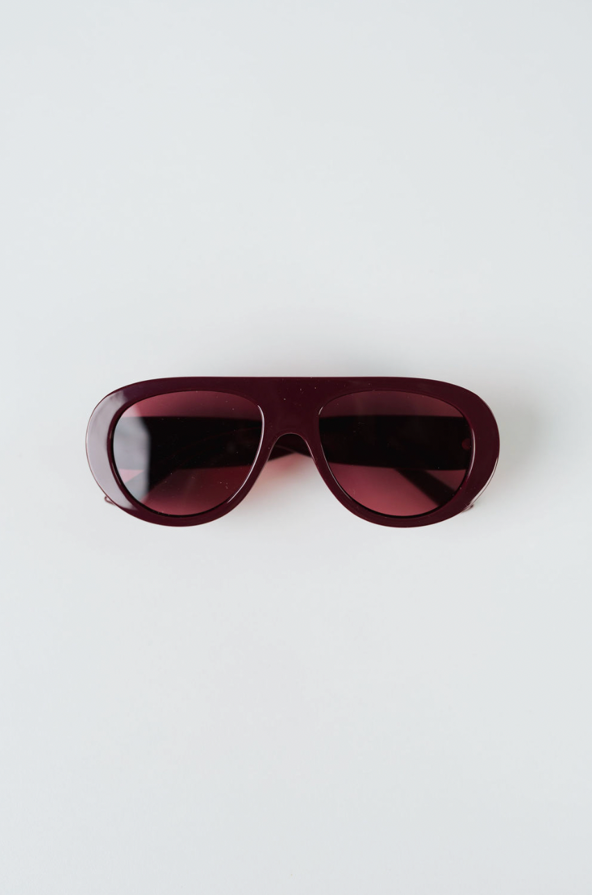 A pair of oversized aviator-style sunglasses with a translucent deep burgundy frame and matching tinted lenses, photographed flat on a light gray background.