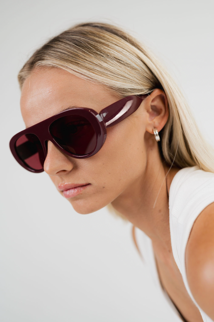 A close-up side profile of a woman with blonde hair wearing oversized aviator-style sunglasses with dark burgundy frames and matching dark lenses, against a plain light gray background.