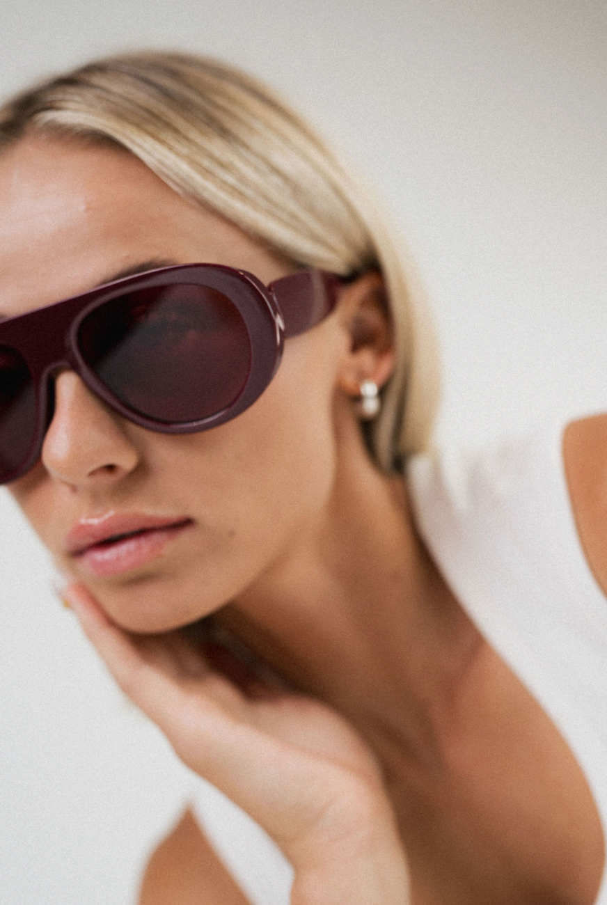 A close-up lifestyle shot of a blonde woman wearing oversized dark burgundy sunglasses and a white top, resting her chin on her hand against a plain light background.