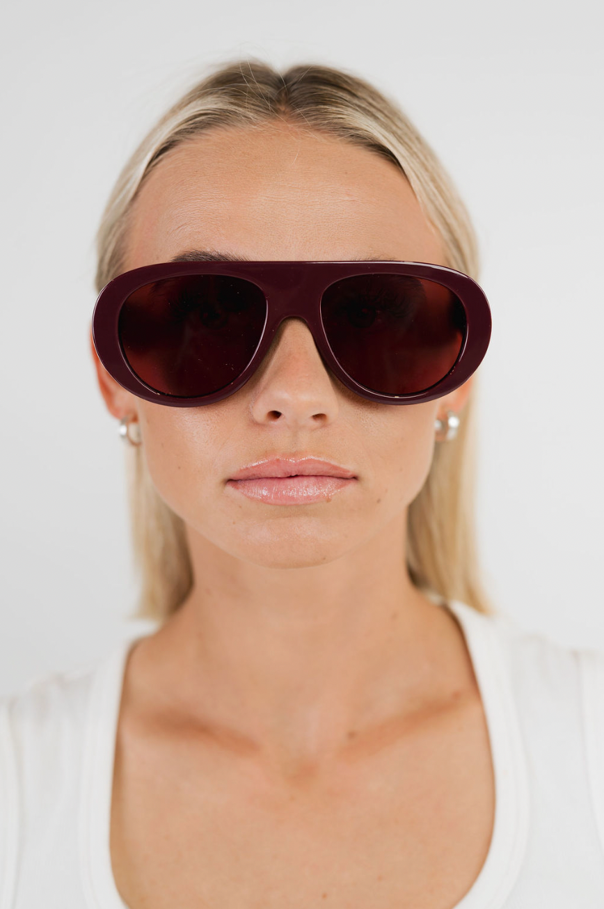 A close-up of a woman with blonde hair wearing oversized aviator-style sunglasses with a dark burgundy frame and matching dark lenses, against a plain light background.