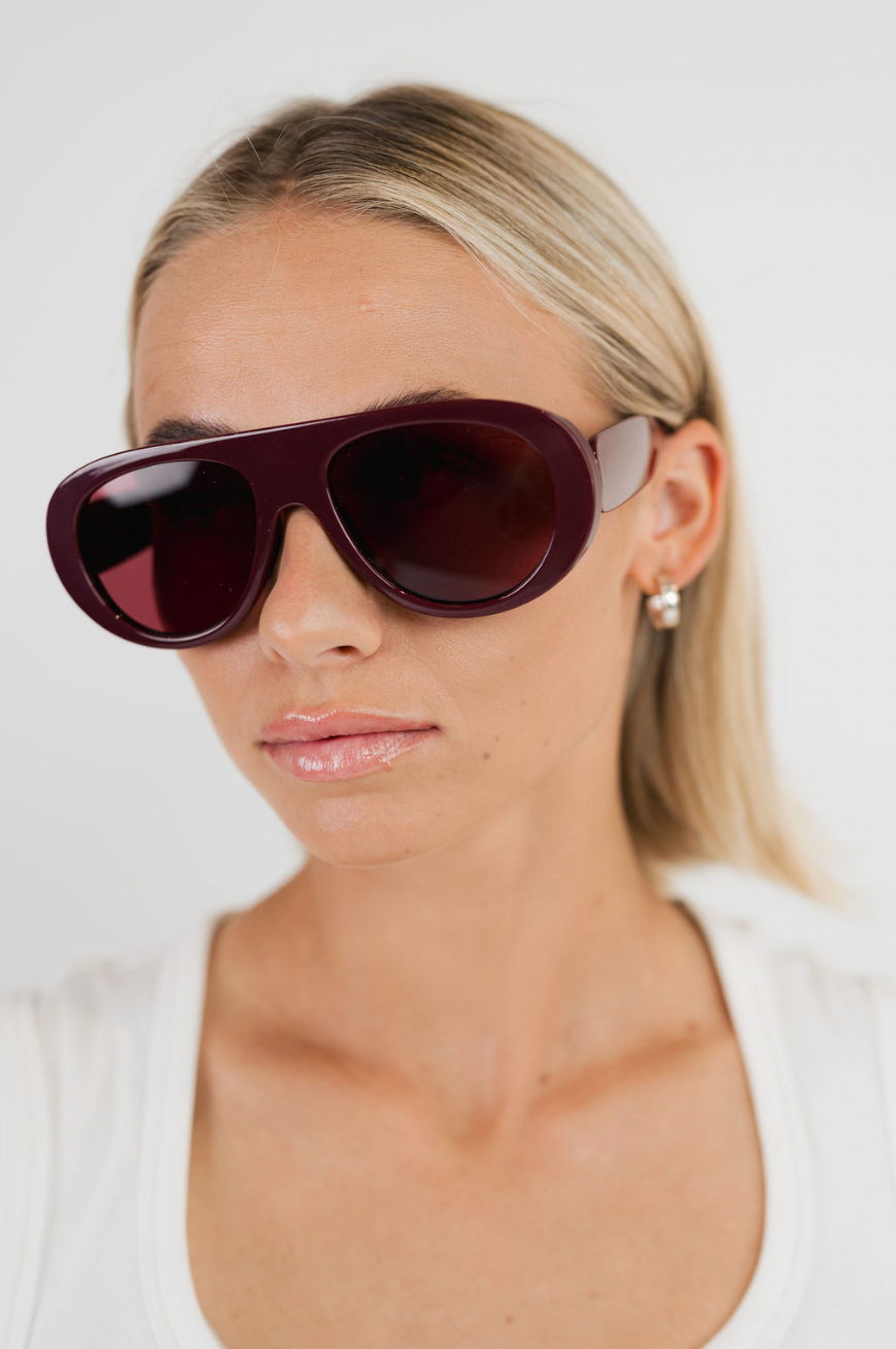 A close-up of a blonde woman wearing oversized aviator-style sunglasses with a deep burgundy frame and dark lenses, paired with a white top against a plain light background.