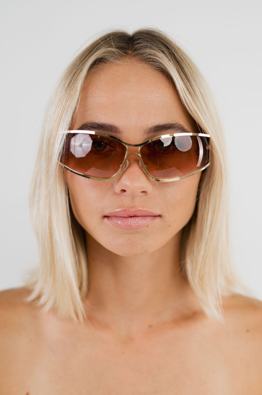 A close-up portrait of a woman with blonde hair wearing oversized square sunglasses with a gold-toned metal frame and brown tinted lenses, set against a plain light gray background.