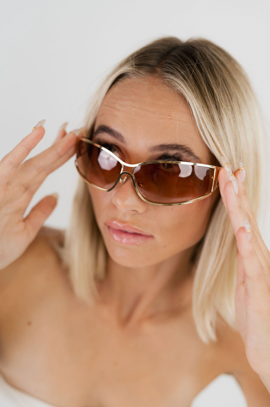 A close-up lifestyle shot of a blonde woman wearing oversized square sunglasses with a gold metal frame and brown lenses, holding them in place with both hands against a clean white background.