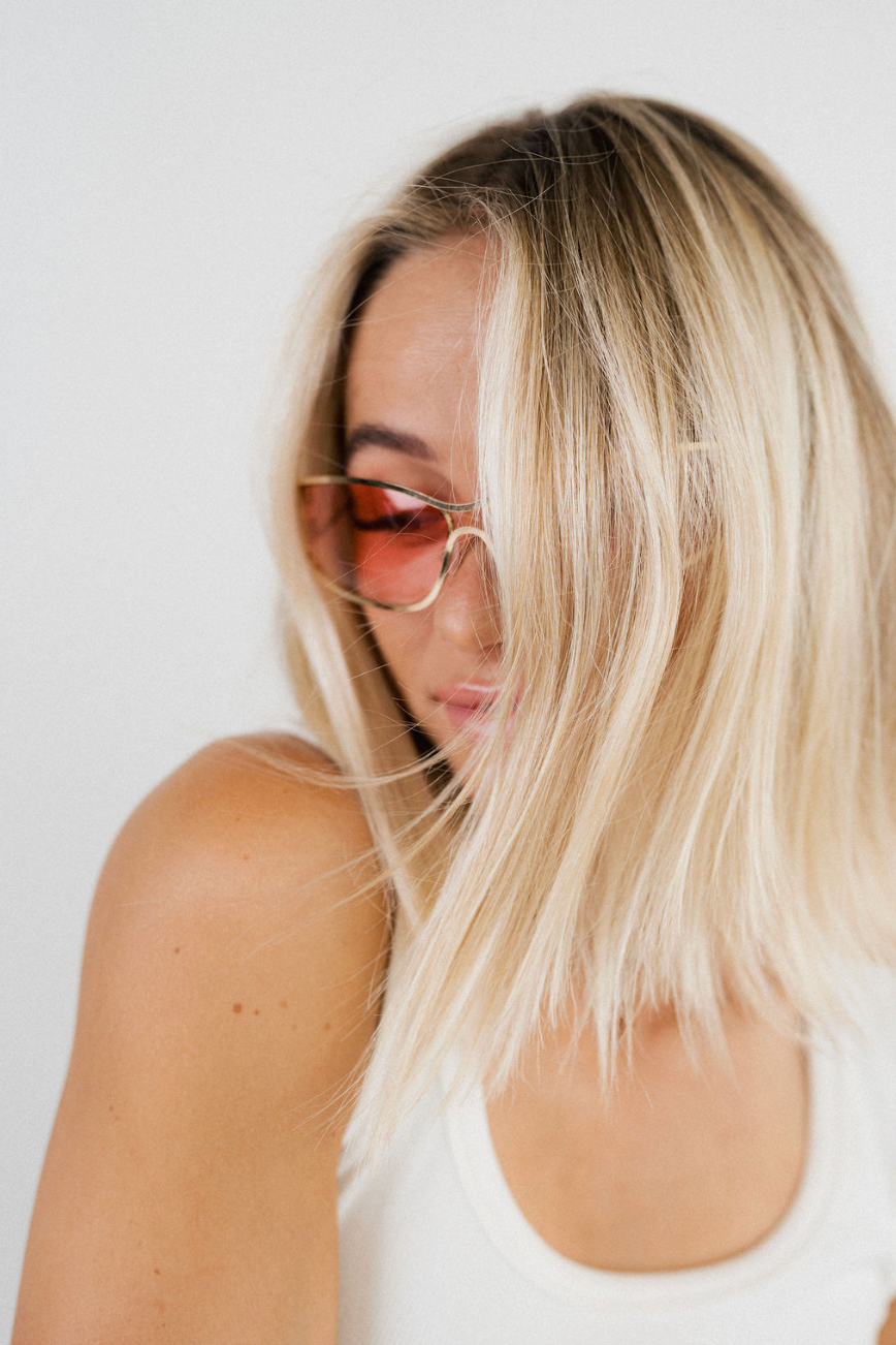 A close-up lifestyle shot of a woman with blonde hair and pink-tinted sunglasses, wearing a white tank top against a plain white background.