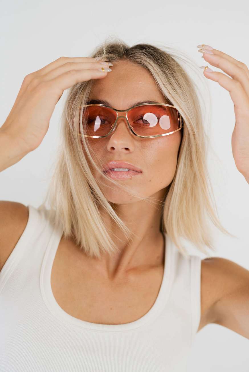 A blonde woman wearing oversized square sunglasses with a gold frame and orange-tinted lenses, posing against a light gray studio background.