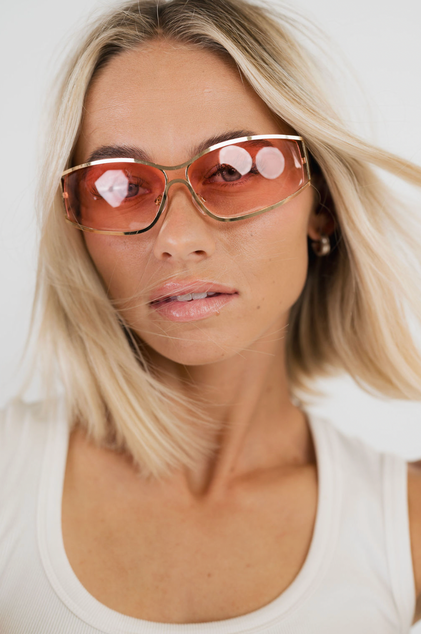 A close-up of a blonde woman wearing oversized square sunglasses with a gold frame and translucent pink-tinted lenses, paired with a white ribbed tank top against a clean white background.