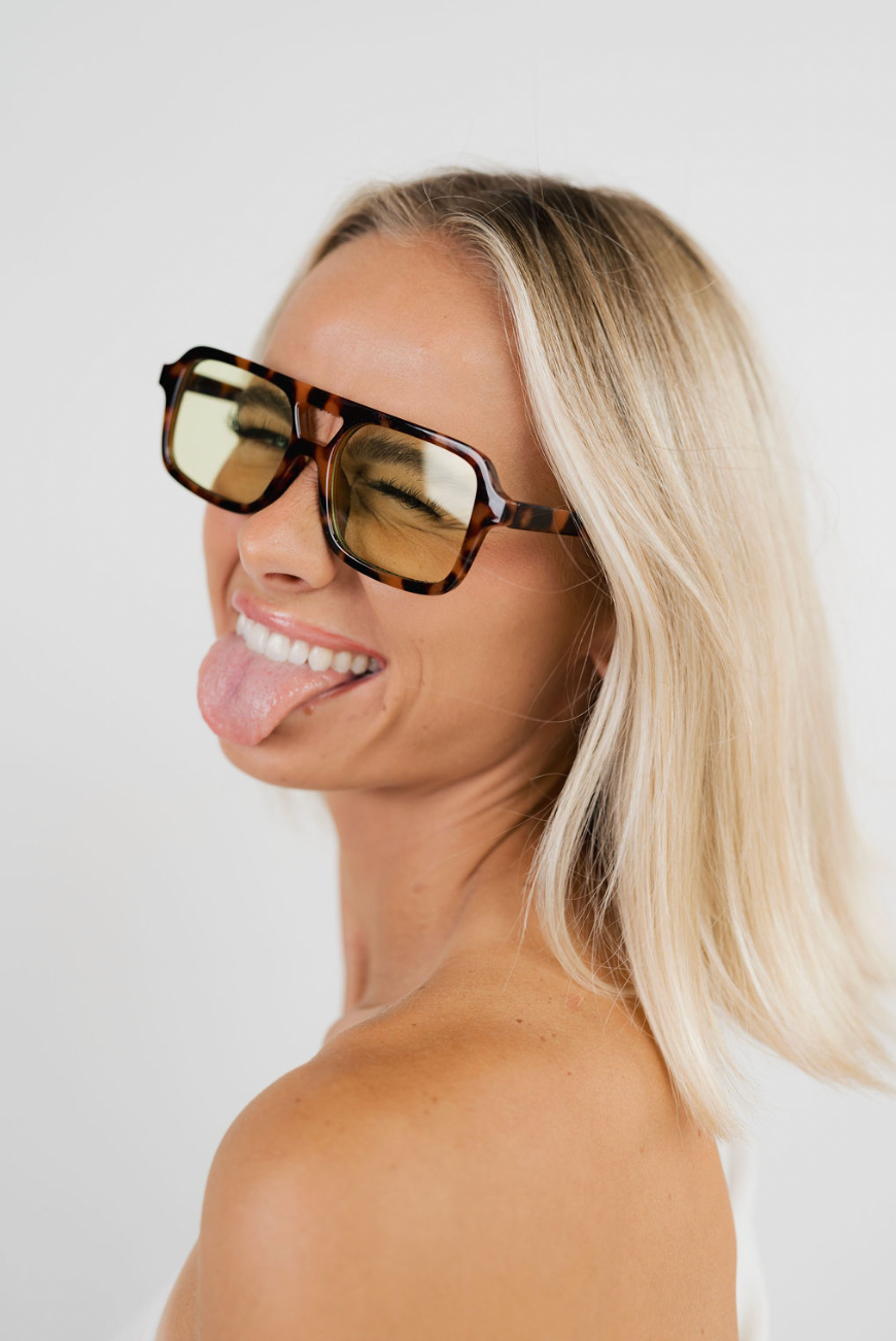 A smiling blonde woman wearing oversized square sunglasses with a tortoiseshell pattern and yellow-tinted lenses, posed against a plain light gray background.