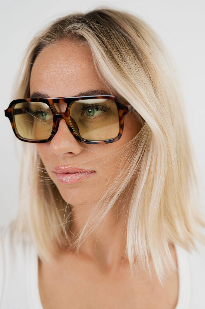 A close-up of a woman with blonde hair wearing oversized square sunglasses with a tortoiseshell pattern and yellow-tinted lenses.
