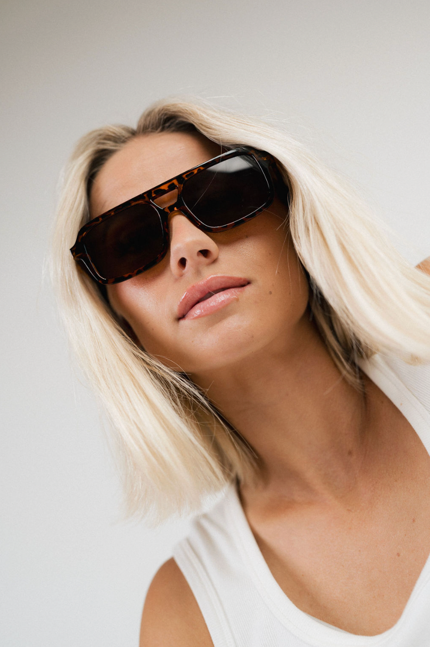 A close-up lifestyle shot of a woman with blonde hair wearing tortoiseshell sunglasses and a white tank top against a plain light gray background.