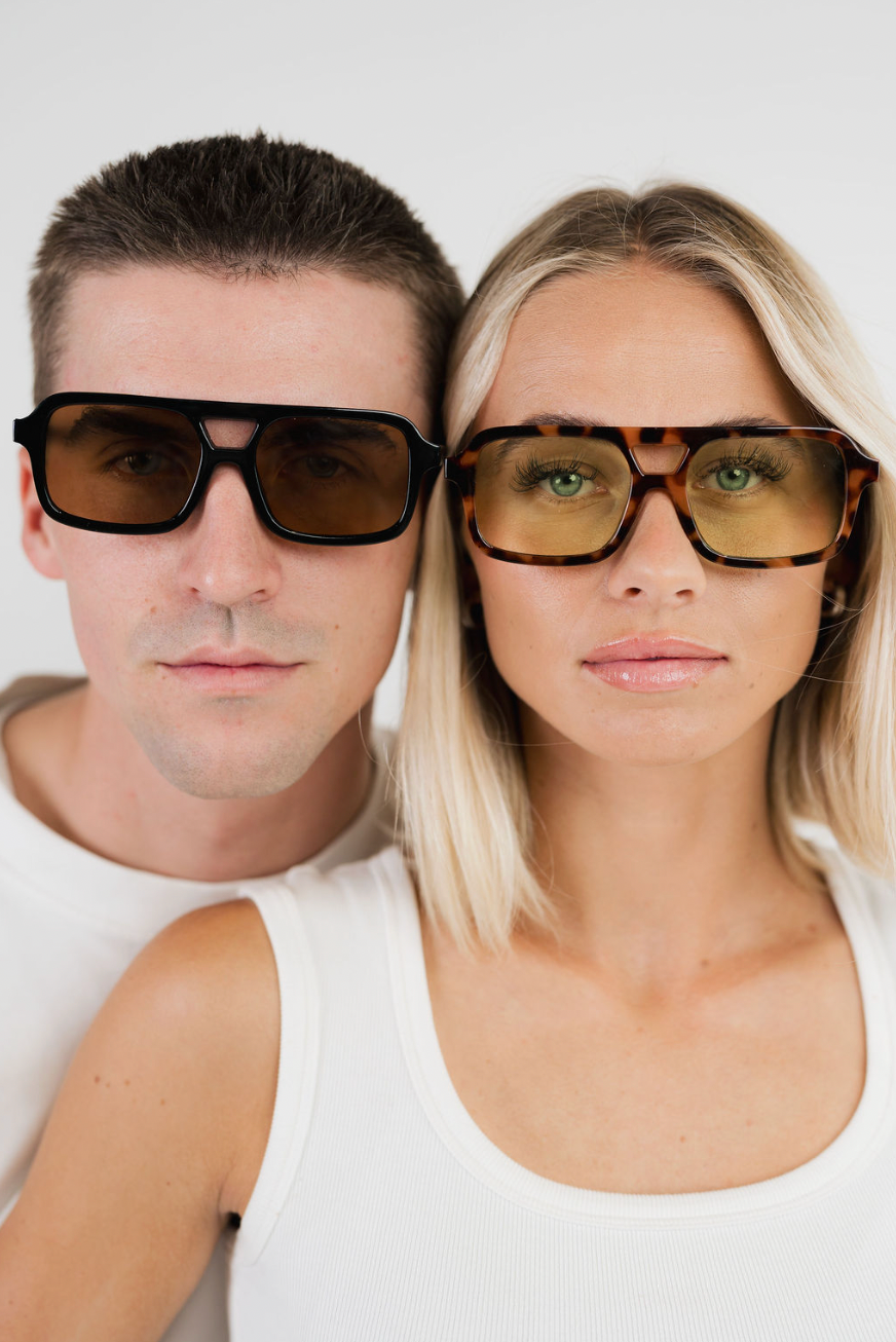 A man and a woman wearing oversized square sunglasses with thick black frames and tinted lenses, posed closely against a plain light background.