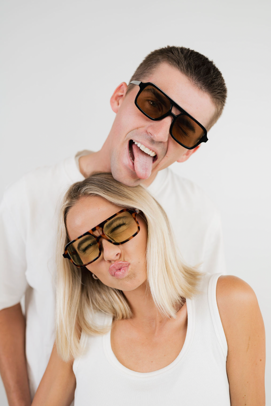 A man and a woman posing together in funny tilted sunglasses. The man has short brown hair and is sticking his tongue out, wearing black sunglasses, while the woman has blonde hair and is puckering her lips, wearing tortoiseshell sunglasses.