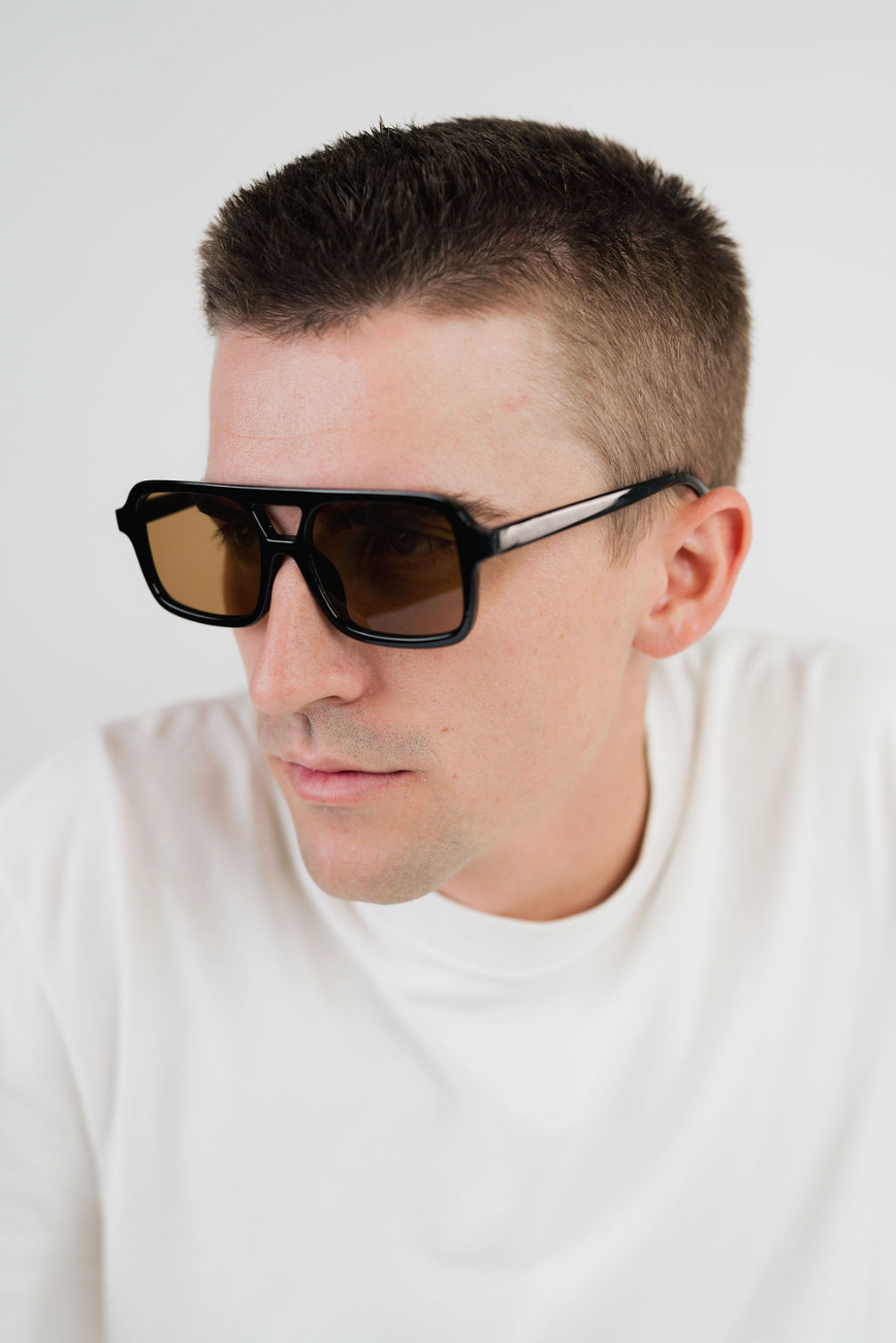 A close-up of a man wearing thick black sunglasses with brown lenses and silver temple tips, paired with a white t-shirt against a plain light background.