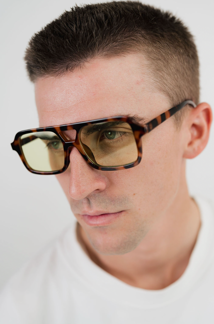 A close-up shot of a man wearing thick, angular tortoiseshell sunglasses with yellow-tinted lenses, set against a plain white background.