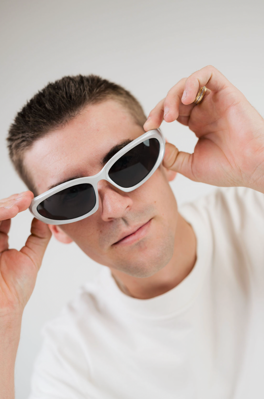 A man wearing white cat-eye sunglasses with dark lenses, holding them in place with both hands against a plain light gray background.