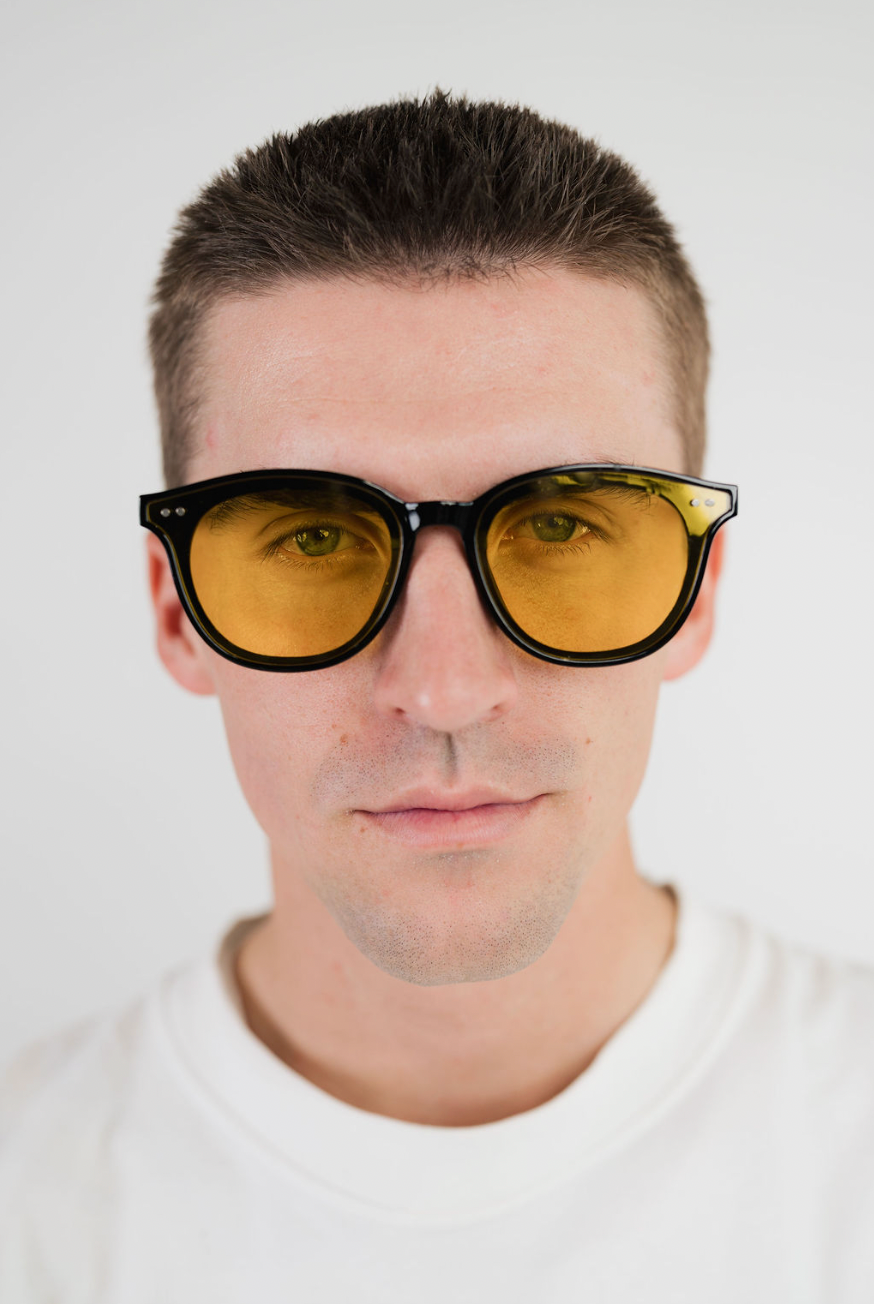 A close-up portrait of a man wearing round sunglasses with black frames and amber-yellow tinted lenses, set against a plain light gray background.