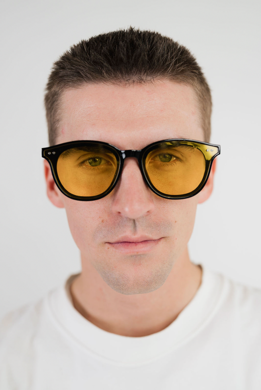 A close-up portrait of a man wearing round sunglasses with black frames and amber-yellow tinted lenses, set against a plain light gray background.