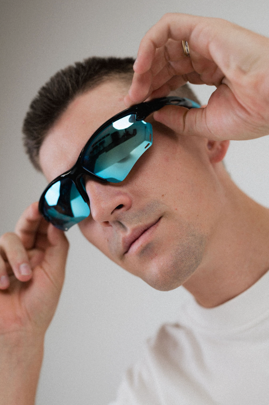 A close-up of a man wearing black wrap-around sunglasses with blue mirrored lenses, holding them in place with both hands against a plain light gray background.