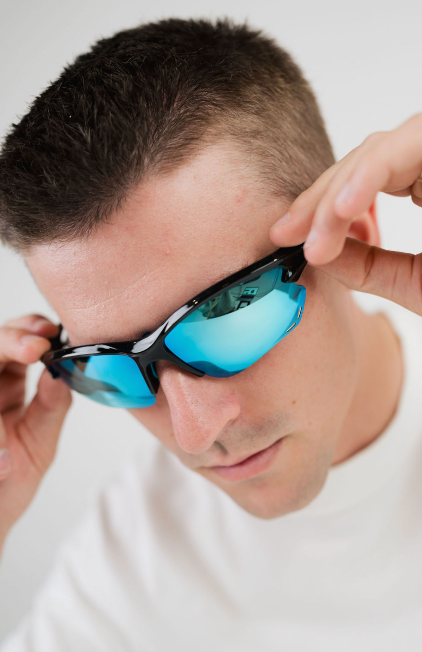 A close-up of a man wearing black sport sunglasses with vibrant blue mirrored lenses, adjusting the frames with both hands against a plain white background.