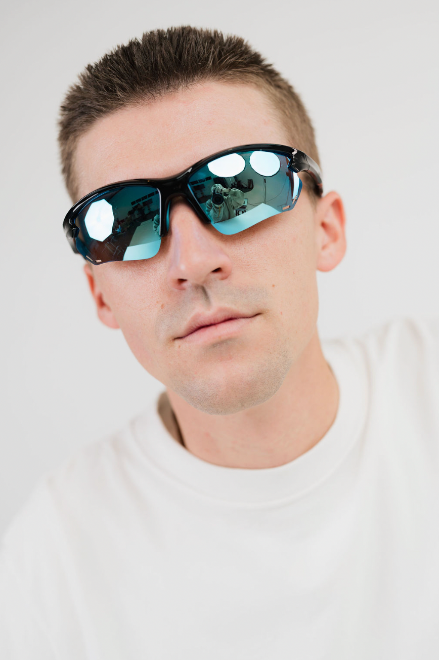 A close-up shot of a man wearing black wrap-around sunglasses with reflective blue-green mirrored lenses, set against a plain light gray background.