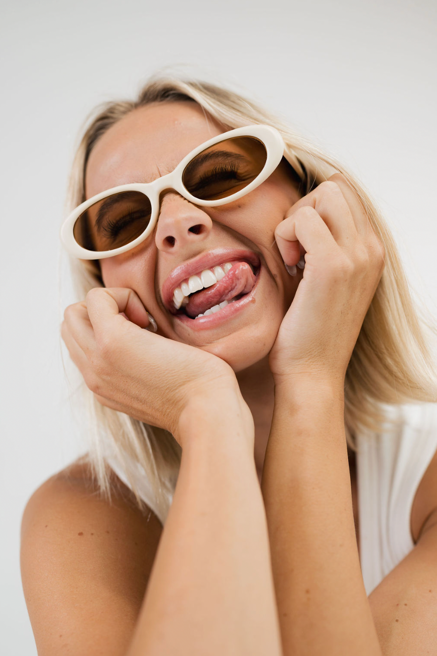A close-up lifestyle shot of a blonde woman wearing chunky, oval-shaped cream-colored sunglasses with dark lenses, posing with her hands on her cheeks against a plain light background.