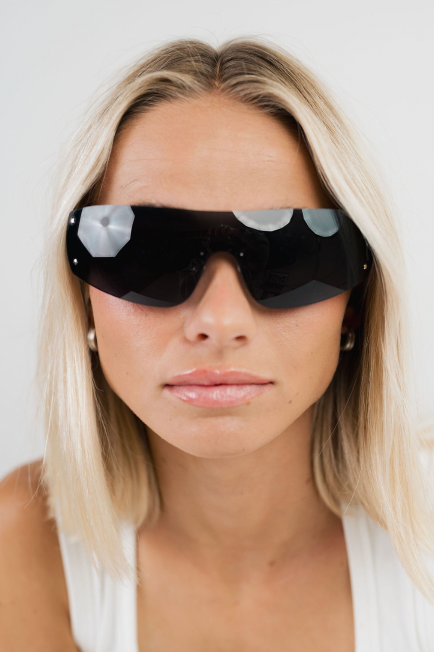 A close-up of a blonde woman wearing futuristic wraparound sunglasses with a glossy black frame and reflective silver geometric lenses, set against a plain light gray background.