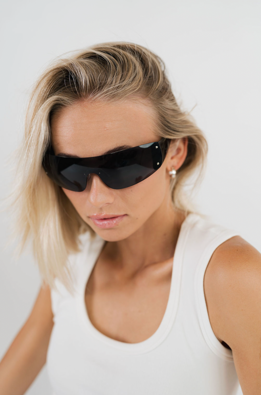 A woman with blonde hair wearing black shield-style sunglasses and a white tank top, photographed against a plain light background.