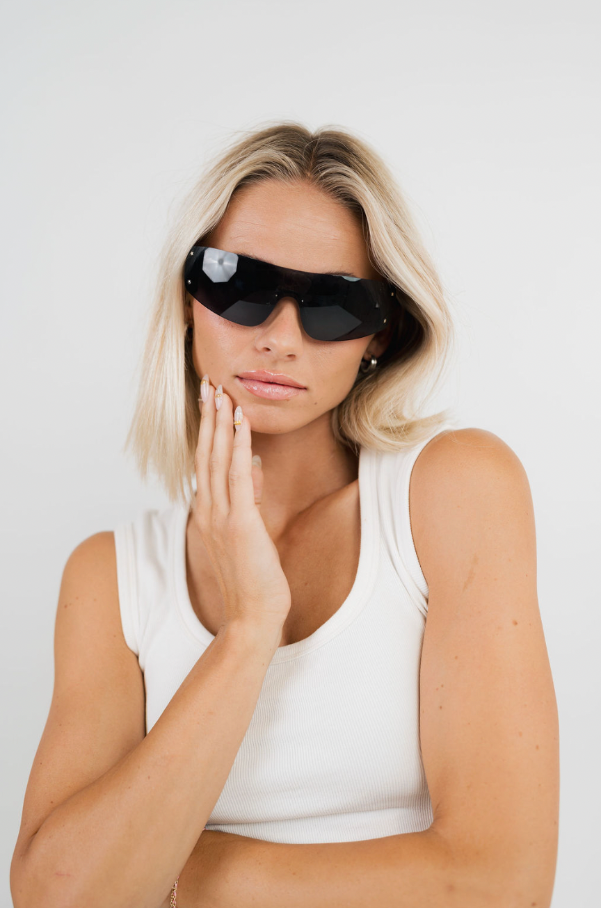 A blonde woman wearing black wrap-style sunglasses and a white tank top, posing with her hand near her face against a plain light background.