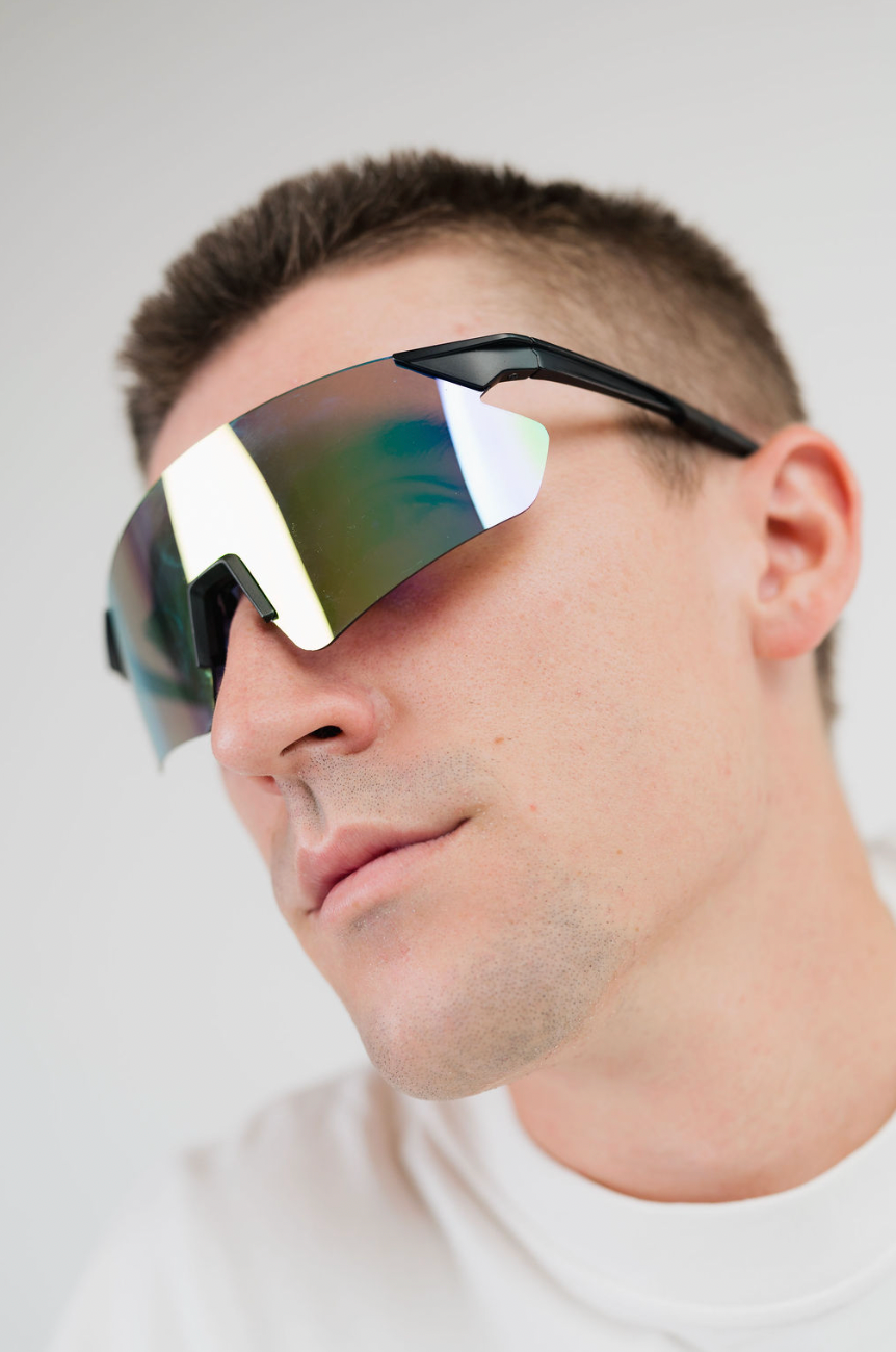 A close-up of a man wearing shield-style sunglasses with a black frame and reflective mirrored lenses that shift from green to gold and silver.