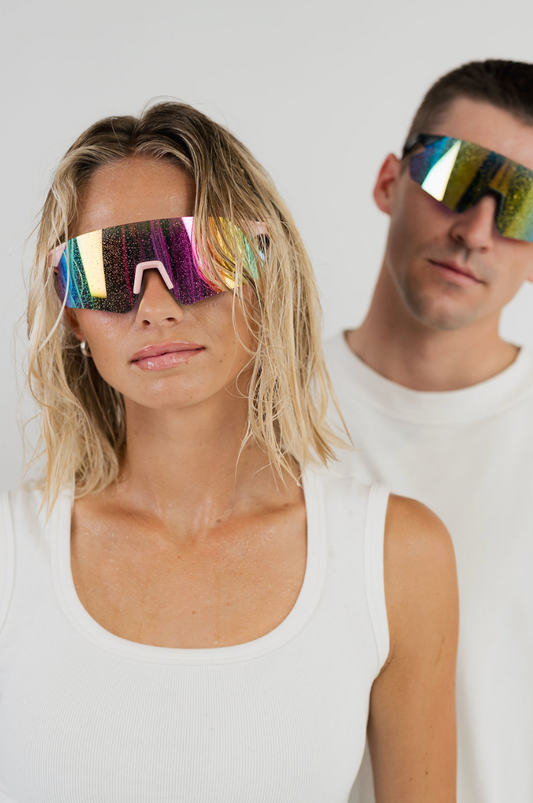 A woman in the foreground and a man behind her, both wearing oversized reflective shield-style sunglasses with a multicolor iridescent finish and white tank tops against a light gray studio background.