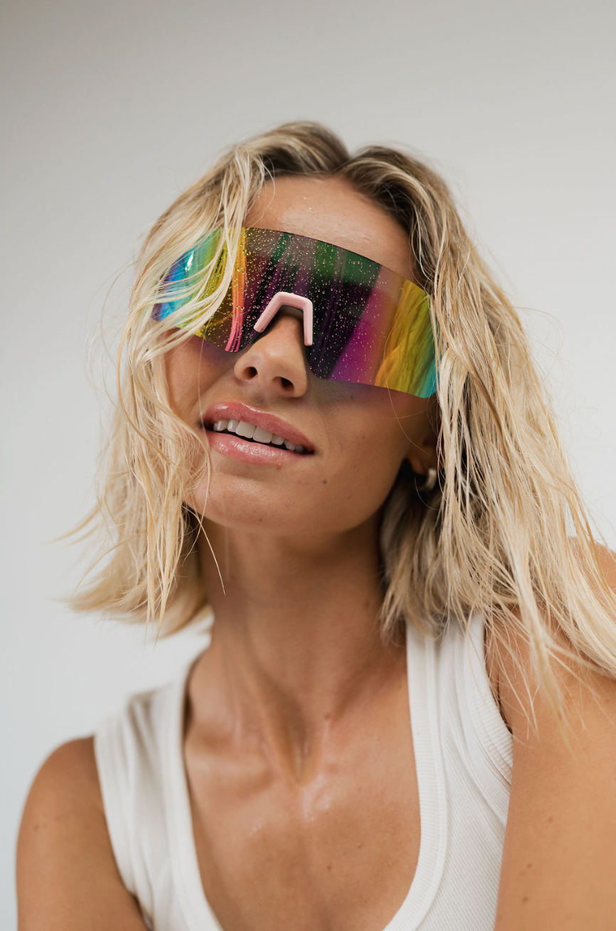 A close-up lifestyle shot of a woman with blonde hair wearing oversized shield sunglasses with a vibrant, multicolored reflective lens and a white ribbed tank top, set against a plain light gray background.