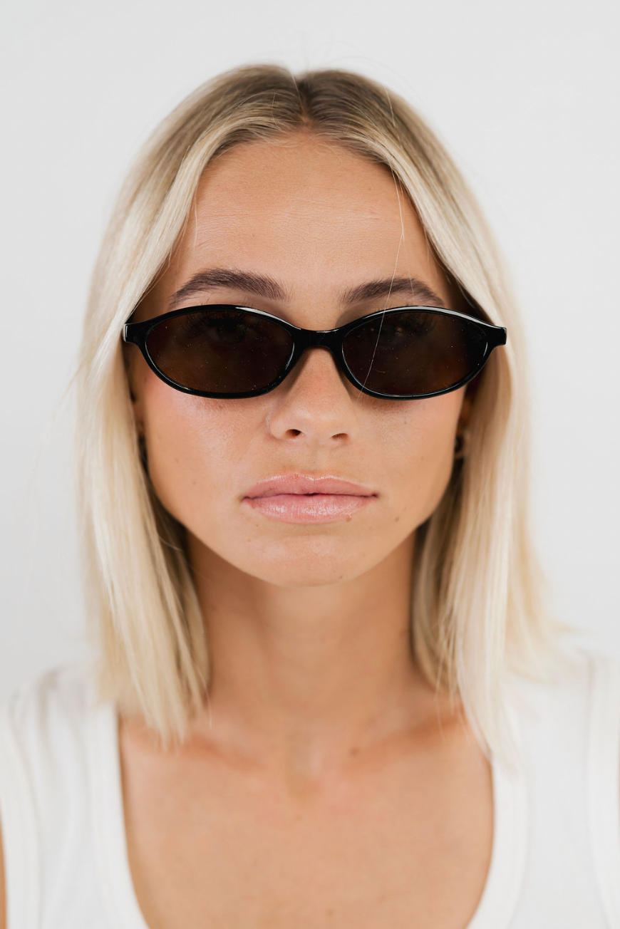 A close-up of a blonde woman wearing black cat-eye sunglasses with dark lenses, set against a plain white background.