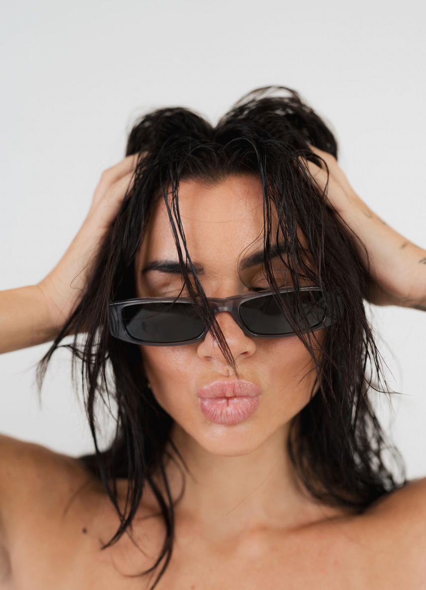 A close-up lifestyle shot of a woman with wet, messy dark hair wearing dark rectangular sunglasses and kissing her lips, set against a plain light gray background.