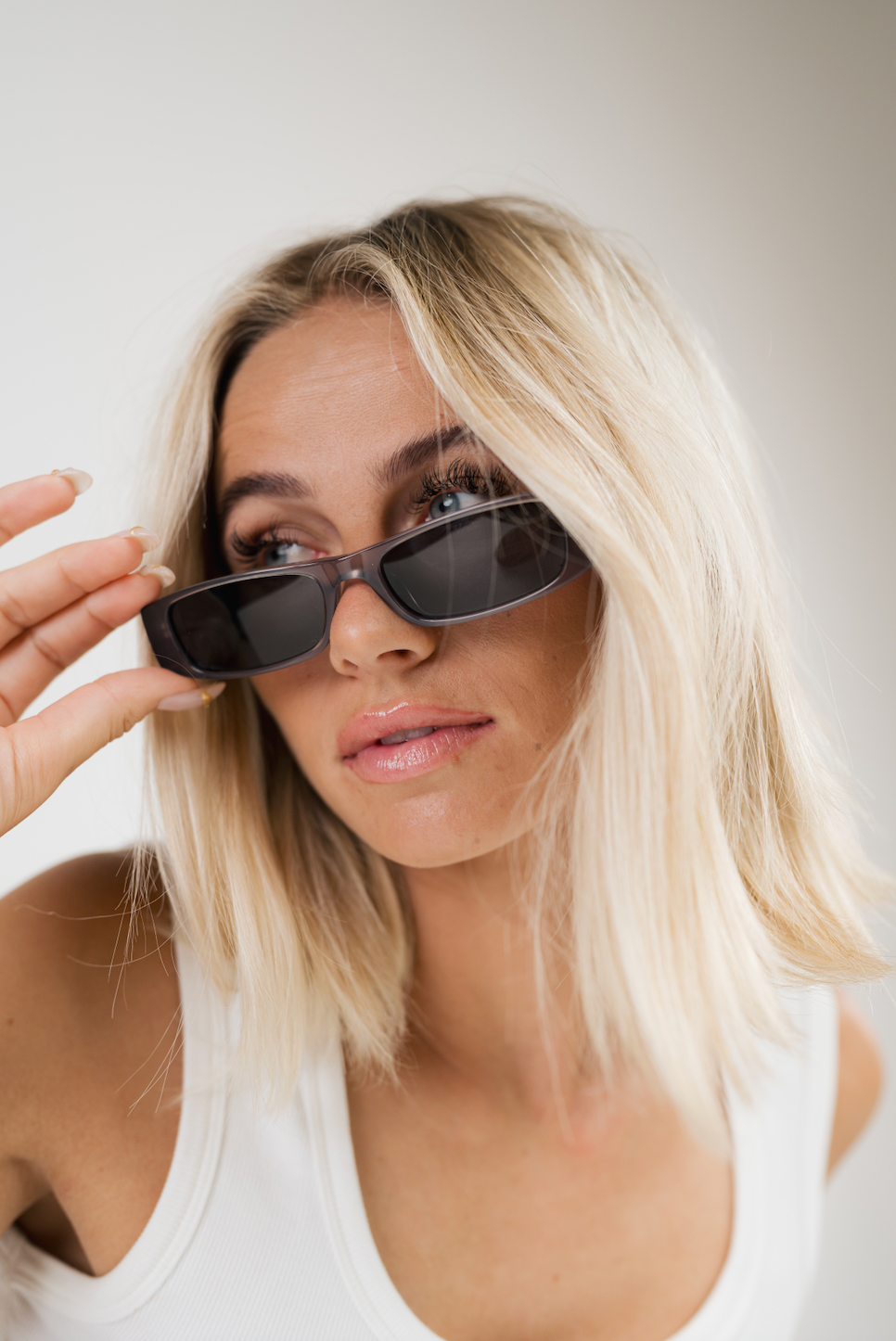 A close-up lifestyle shot of a blonde woman wearing dark rectangular sunglasses and a white tank top, holding one of the sunglasses with her hand against a plain light gray background.