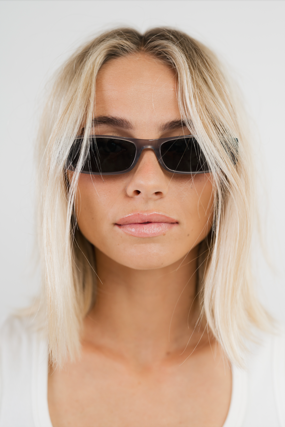 A close-up portrait of a woman with blonde hair wearing dark rectangular sunglasses and a white top against a plain light background.